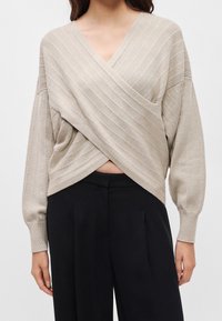 Femme portant un pull en maille côtelée beige avec un devant croisé et un pantalon noir taille haute à jambes larges.