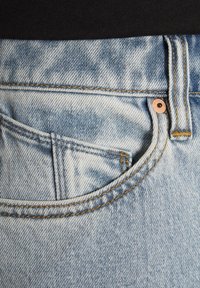 Jeans de mezclilla azul claro con un bolsillo frontal cosido, botón de cobre y costuras amarillas a lo largo de las costuras. La textura parece ligeramente desgastada y desvanecida.