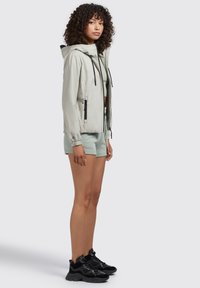 Lichtbeige windbreaker met een capuchon, voorritsen en elastische manchetten. Gecombineerd met mintgroene shorts en zwarte sneakers. Soepele textuur.