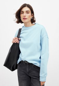 Sudadera azul clara con cuello redondo y hombros caídos, combinada con jeans oscuros y un bolso tote de cuero negro.