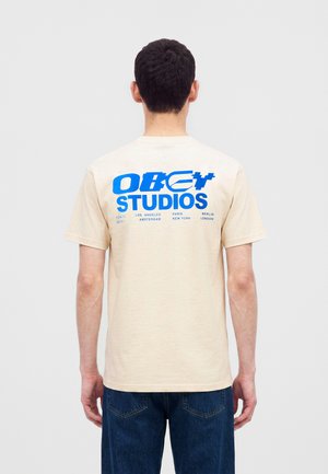 Muž s krátkymi tmavými vlasmi, ktorý nosí béžové tričko s modrým textom "OBEY STUDIOS" a názvami miest na chrbte, v kombinácii s tmavomodrými džínsami.