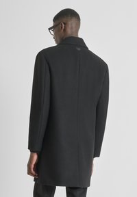 Cappotto in misto lana nero con colletto standard, taglio dritto e maniche lunghe. Presenta uno spacco dietro e dettagli di design minimalisti.