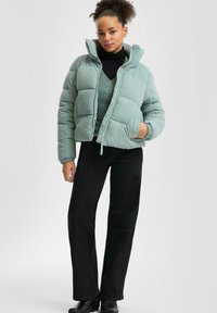 Padded light teal jacket with a high collar, zip closure, and ribbed cuffs, worn over a black sweater and paired with black pants.