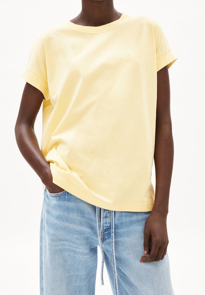 Yellow cotton t-shirt with rolled sleeves, crew neck, and small logo detail on the chest, paired with light blue denim pants.