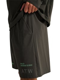 Persoon draagt donkere, gestructureerde korte broek met zijzak, hand tilt bijpassende top op, groene tekst “HALO 196819721981” en zwarte letters “ATW” zichtbaar.