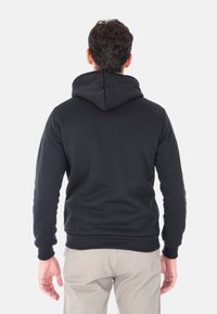 Uomo con capelli scuri che indossa una felpa nera con cappuccio e pantaloni beige, in piedi con le spalle rivolte verso un fondo bianco puro.