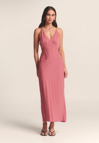 Ilga, prigludusi suknelė iš švelnios rožinės medžiagos, su gilia V formos iškirpte, plonomis petnešėlėmis ir suvertu krūtinės detalės audiniu, siekianti vidurio blauzdos.