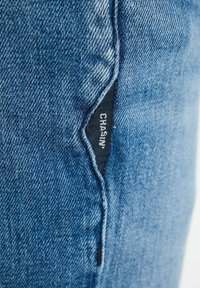 Modrá denimska tkanina s bočnou vreckom a čiernym štítkom, na ktorom je napísané "CHASIN'". Textúra zobrazuje klasické denimové tkanie s miernym vyblednutím.