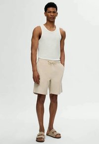Top de alças branco com textura canelada, calções beje com cordão na cintura e chinelos de slide castanhos claros. Tecido suave, design casual, adequado para clima quente.