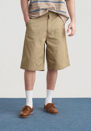 Pantaloncini beige in cotone con taglio dritto. Presentano tasche anteriori e chiusura con bottone. Abbinati a scarpe marroni e calzini bianchi.