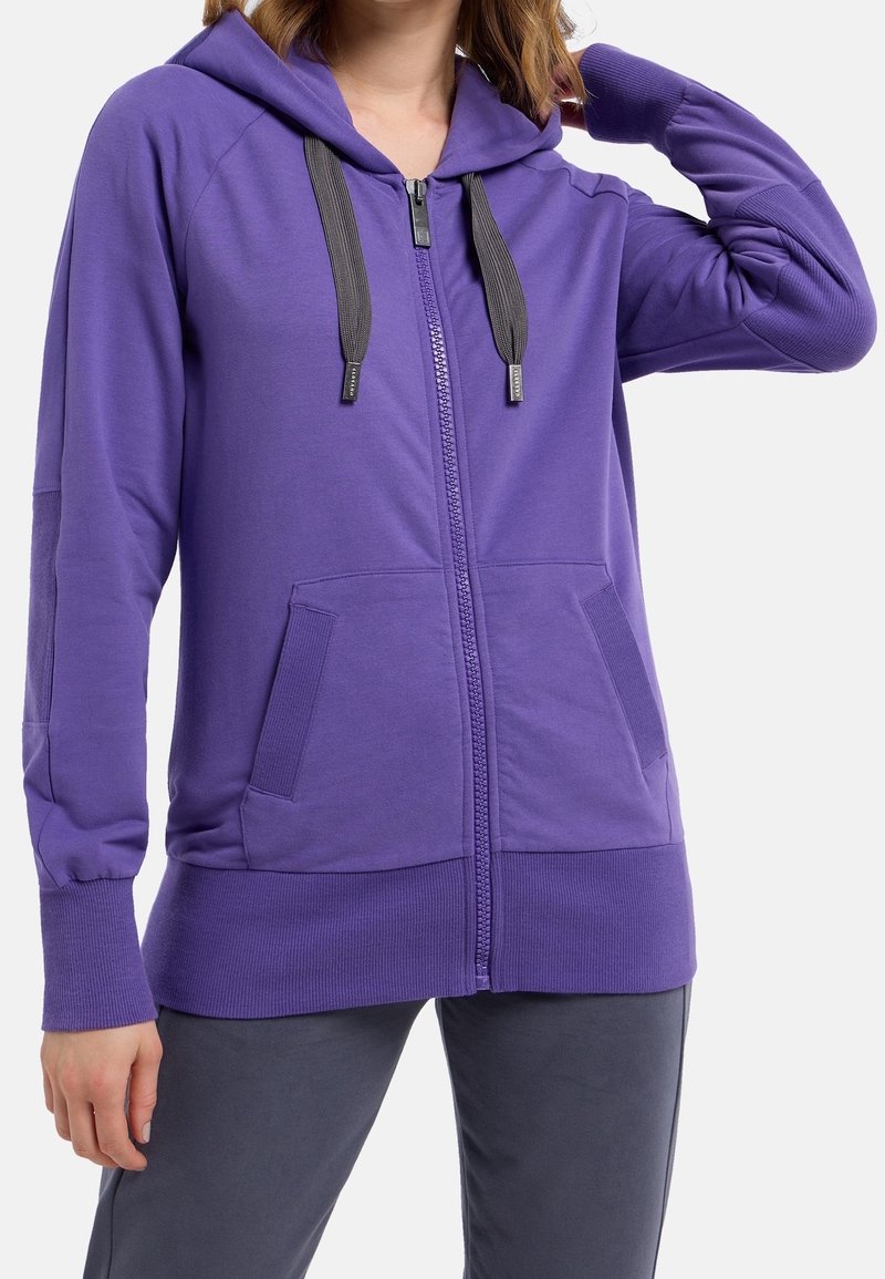 Femme portant un sweat à capuche violet zippé avec des cordons noirs et des poches avant, associé à un pantalon gris foncé, main levée près de la tête.