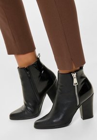 Svarta läderankelstövlar med spetsig tå och dragkedjedetalj på utsidan. Har en tjock klack och en slät yta.