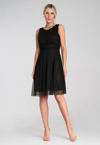 Schwarzes Kleid mit tailliertem Oberteil, runder Halslinie und einem fließenden Rock. Transparentes Overlay sorgt für Textur. Dazu werden hautfarbene Ankle-Strap-Absätze getragen.