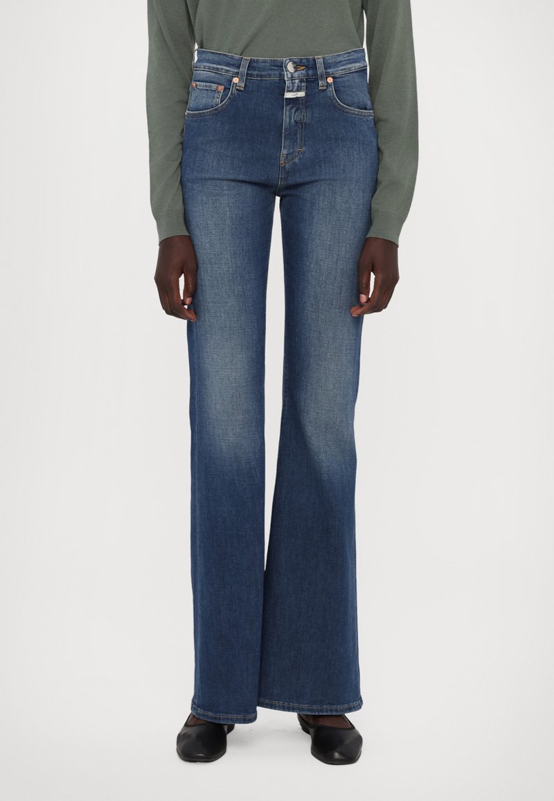 Jeans de mezclilla azul acampanados con una cintura alta, que cuentan con cinco bolsillos, costuras en naranja y un sutil detalle de lavado. Acompañados de zapatos negros.