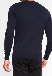 Pull bleu marine à manches longues avec poignets et ourlet côtelés ; texture en tricot lisse ; vue de dos montrant un subtil détail de couture horizontale.