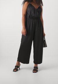 Mono de satén negro con escote en V, tirantes ajustables, cintura elástica, perneras anchas y una textura suave. Sandalias de tacón negro.