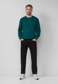 Felpa verde acqua con scollatura tonda e logo sul petto, abbinata a pantaloni neri. Il modello sta in piedi con le mani in tasca, indossando scarpe da ginnastica chiare.