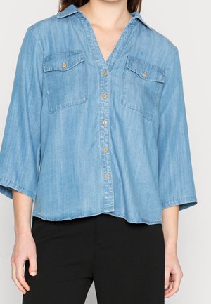 Kvinde iført en lyseblå denimskjorte med knapper, to brystlommer og trekvartærmer, kombineret med sorte bukser.