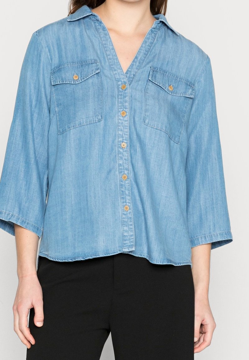 Femme portant une chemise en denim bleu clair à boutons, avec deux poches poitrine et des manches trois-quarts, associée à un pantalon noir.