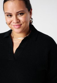 Lachend persoon met donker haar naar achteren gebonden, zilveren oorbellen in de vorm van ringen, gelaagde zilveren kettingen en een zwart bovenstuk met kraag.