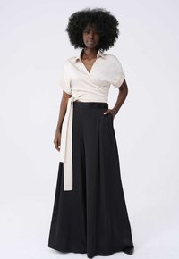 Beige wikkelblouse met korte mouwen en een strikdetail, gecombineerd met hooggesneden zwarte wijde broek met een gladde textuur en zakken.