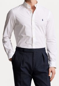 Witte overhemd met knopen en een klein blauw geborduurd logo, gecombineerd met marineblauwe pantalon met krijtstrepen, een vlakke voorkant en een riem.