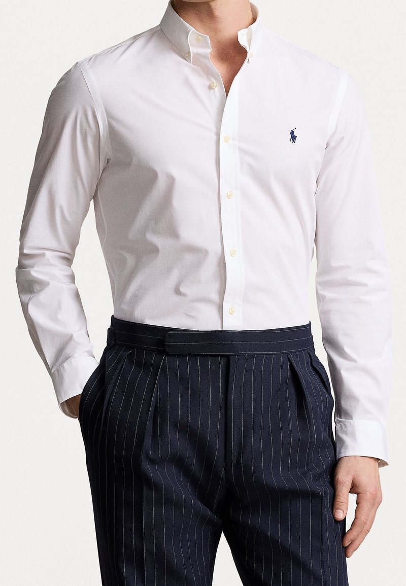 Witte overhemd met knopen en een klein blauw geborduurd logo, gecombineerd met marineblauwe pantalon met krijtstrepen, een vlakke voorkant en een riem.