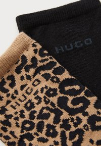 Deux paires de chaussettes : une noire unie, l'autre beige avec un motif léopard noir. Matériel en tricot doux avec des poignets côtelés. Nom de la marque "HUGO" en bleu.