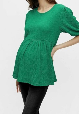 Vrouw die een getextureerde groene zwangerschaps top met pofmouwen en zwarte broek draagt, poserend met één hand op haar heup en de andere langs haar zijde.