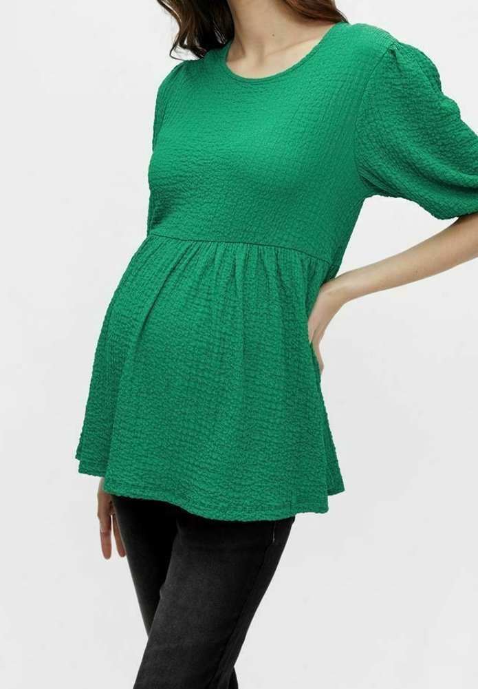 Femme portant un haut de maternité vert texturé avec des manches bouffantes et un pantalon noir, posant avec une main sur la hanche et l'autre le long du corps.