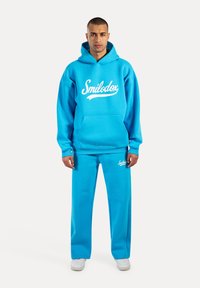 Sudadera azul y pantalones a juego hechos de tela suave con un bolsillo frontal. Logo de "Smilodox" en blanco en ambas piezas.