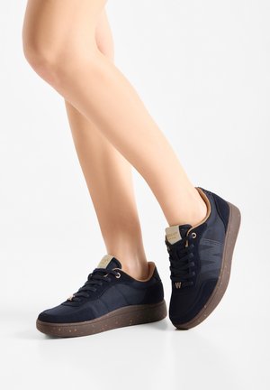 Person iført marineblå sneakers med brune plettede såler og beige indvendig for på hvid baggrund.