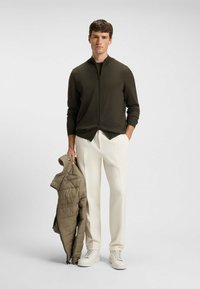 Donkergroene ritsjack gemaakt van gebreid stof, witte wijde broek, en lichtbeige pufferjack in de hand. Witte sneakers.