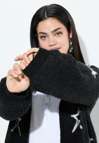 Un cardigan en tricot noir avec une texture épaisse, des poignets côtelés et des motifs d'étoiles blanches. Porté par-dessus une chemise blanche à col.