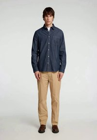 Camisa azul-escura de botão com um bolso, combinada com calças caqui claras e sapatos castanhos, num modelo em pé contra um fundo simples.
