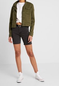 Chaqueta verde oliva recortada con dos bolsillos frontales, combinada con pantalones cortos de mezclilla negros y zapatillas altas blancas sobre un fondo neutro.