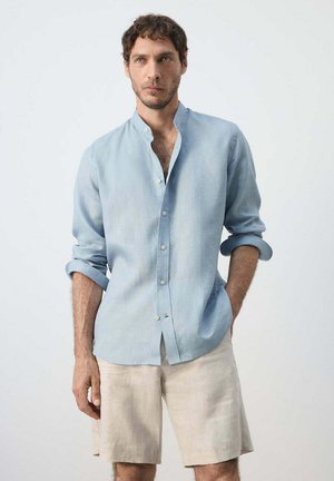 Hombre con cabello corto y rizado y barba clara, que lleva una camisa azul clara de manga larga y pantalones cortos beige, con una mano en el bolsillo.