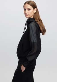Sudadera negra con una textura suave, presenta un cordón y paneles transparentes negros en las mangas. Modelo vistiendo pantalones negros a juego.