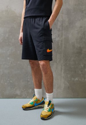 Persoon draagt zwarte cargoshorts met een oranje Nike-logo, witte sportsokken en gele en blauwe Nike-sneakers, staand op een grijze vloer.
