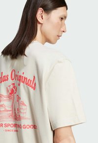Bežinė medvilninė marškinėliai su raudonu spaudiniu, vaizduojančiu asmenį, dirbantį su batais, ir tekstu "Adidas Originals" bei "Sporto prekėms".