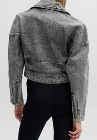 Grå denimjacka i cropped-modell med en strukturerad krage, synlig sömnad och dragkedjor vid ärmarna. Tyget har ett blekt, texturerat utseende.