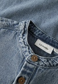 Gaiši zila džinsa krekls ar apaļu apkakli, divām brūnām pogām un birku ar uzrakstu "Lil'Atelier". Audums ar faktūru un nelielām variācijām.