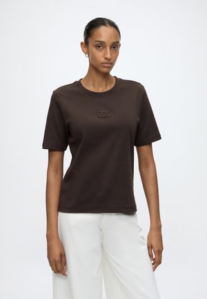 Kvinde med trukket hår bærer en mørkebrun t-shirt med korte ærmer med et lille broderet logo og hvide bukser med brede ben.