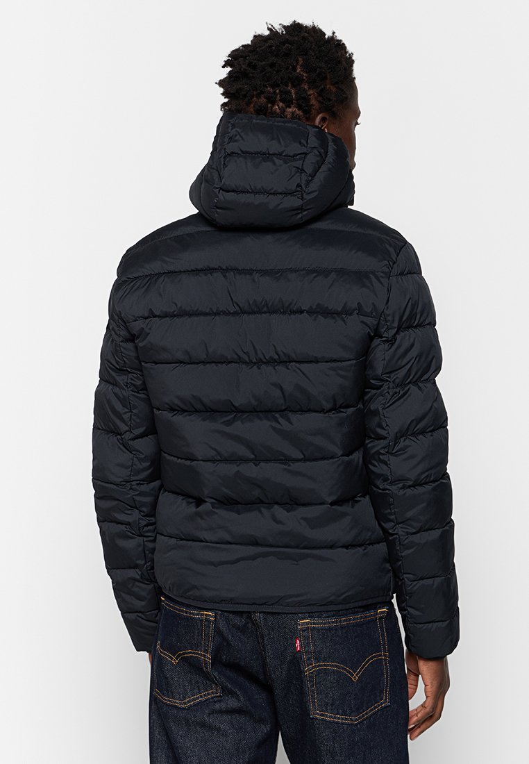 Mężczyzna w czarnej pikowanej kurtce z kapturem i ciemnoniebieskich dżinsach, stojący tyłem na jasnym, jednolitym tle.