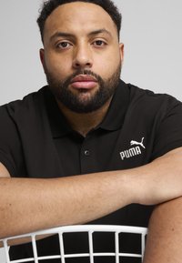 Čierne polo tričko s golierom, na ľavej hrudi s bielym logom Puma. Model má bradu a opiera sa o biely drôtený koš.