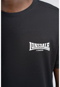 Sort kortærmet t-shirt lavet af bomuld, med en hvid "LONSDALE LONDON" logo på venstre brystområde. Glat tekstur, rund halsudskæring.