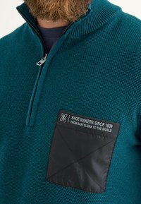 Pull-over teal avec un col zippé, tricot texturé, et une patch en tissu noir avec du texte blanc : "CORDONNIERS DEPUIS 1939 DE BARCELONE AU MONDE."
