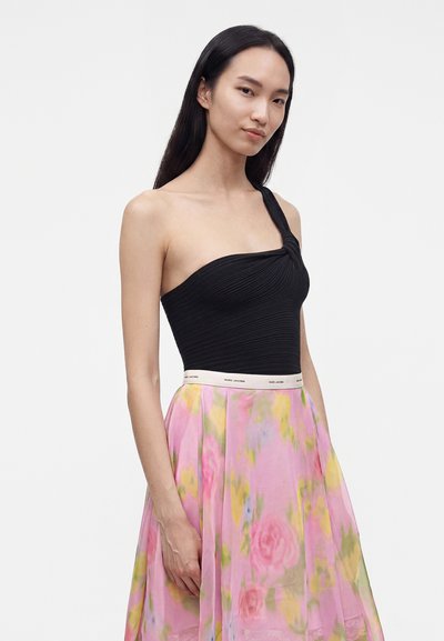 Femme portant un haut noir asymétrique et une jupe rose à fleurs avec une ceinture blanche Marc Jacobs, debout devant un fond uni.