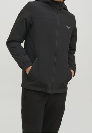 Osoba ubrana w czarną bluzę z kapturem zapinaną na zamek i czarne spodnie, z rękami w kieszeniach bluzy, na jasnoszarym tle.