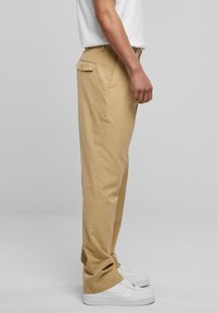 Mann i beige bukser med en knappet baklomme og hvite joggesko, som står på skrå mot en ensfarget lysegrå bakgrunn.